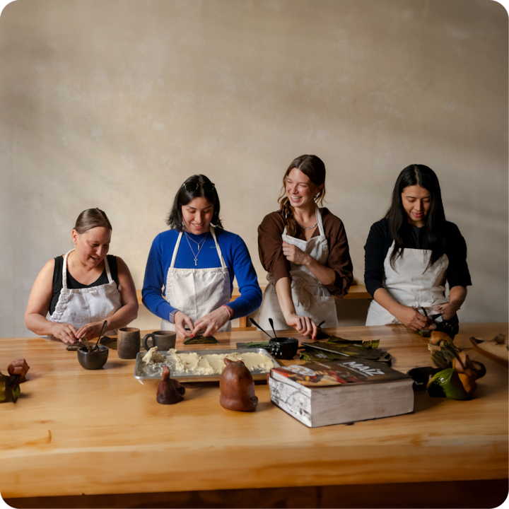 Un groupe vêtu de tabliers est rassemblé autour d'une table en bois. Ses membres façonnent de la pâte et enveloppent des ingrédients dans des feuilles dans le cadre d'un cours pratique de cuisine.