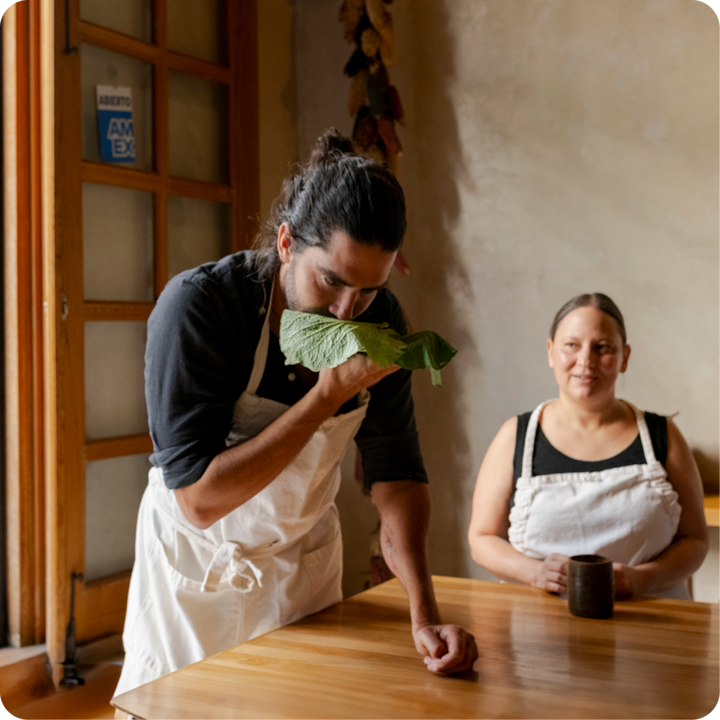Un enseignant de cuisine tient une grande feuille verte près de son visage, debout à une table en bois, tandis qu'une participante assise à proximité portant un tablier blanc tient une tasse.