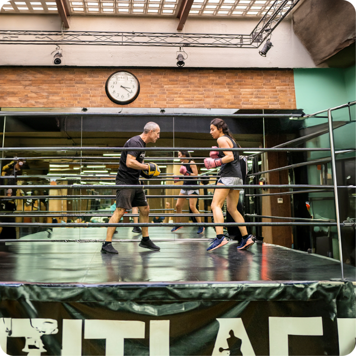 Deux personnes pratiquent la boxe dans un ring avec des miroirs muraux, l'une tenant des pattes d'ours et l'autre portant des gants roses, sous une horloge sur un mur de briques.