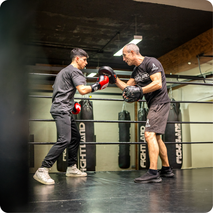 Un coach de fitness tient des pattes d'ours pendant qu'une participante pratique des techniques de boxe dans une salle de sport avec des sacs de boxe suspendus en arrière-plan.