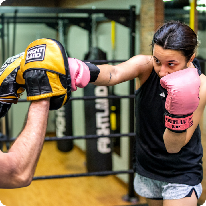 Une personne portant des gants de boxe roses pratique des techniques de frappe avec un entraîneur dans une salle de sport équipée de sacs de boxe et d'un sol amortissant.