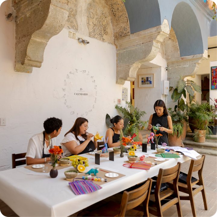 Un groupe de personnes est assis à une table ornée de fournitures colorées et de fleurs fraîches dans un espace lumineux et artistique doté d'une architecture voûtée en pierre.