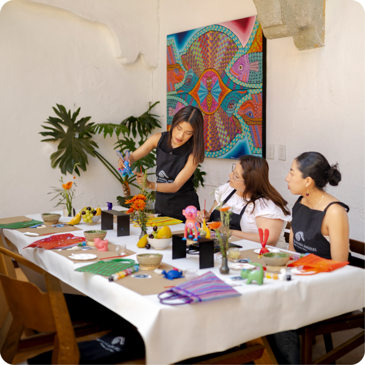 Des participants à un atelier d'artisanat coloré sont assis à une table remplie de fournitures artistiques, d'objets faits main et d'articles décoratifs aux couleurs vives, avec un tableau aux motifs lumineux et des plantes vertes luxuriantes en arrière-plan.