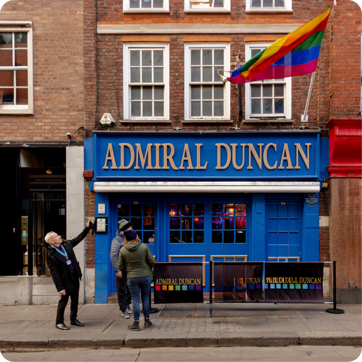 Extérieur du pub Admiral Duncan à Londres avec un drapeau arc-en-ciel flottant au-dessus de l'entrée. Trois personnes se tiennent à l'extérieur, dont une pointant du doigt le bâtiment.