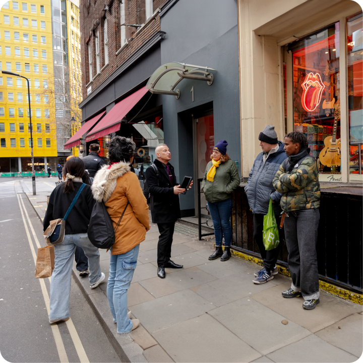 Un guide touristique tenant une tablette parle à un petit groupe de personnes debout sur un trottoir devant un magasin avec un néon du logo des Rolling Stones dans la vitrine. Un auvent rouge et un bâtiment jaune sont visibles en arrière-plan.