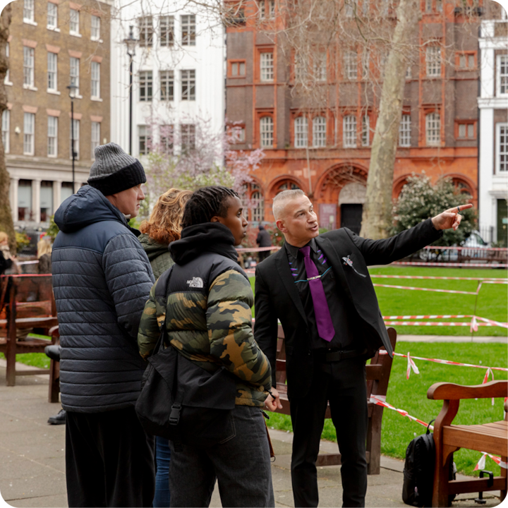 Un guide touristique portant un costume et une cravate violette discute avec un petit groupe en montrant de la main un bâtiment historique entouré de verdure.