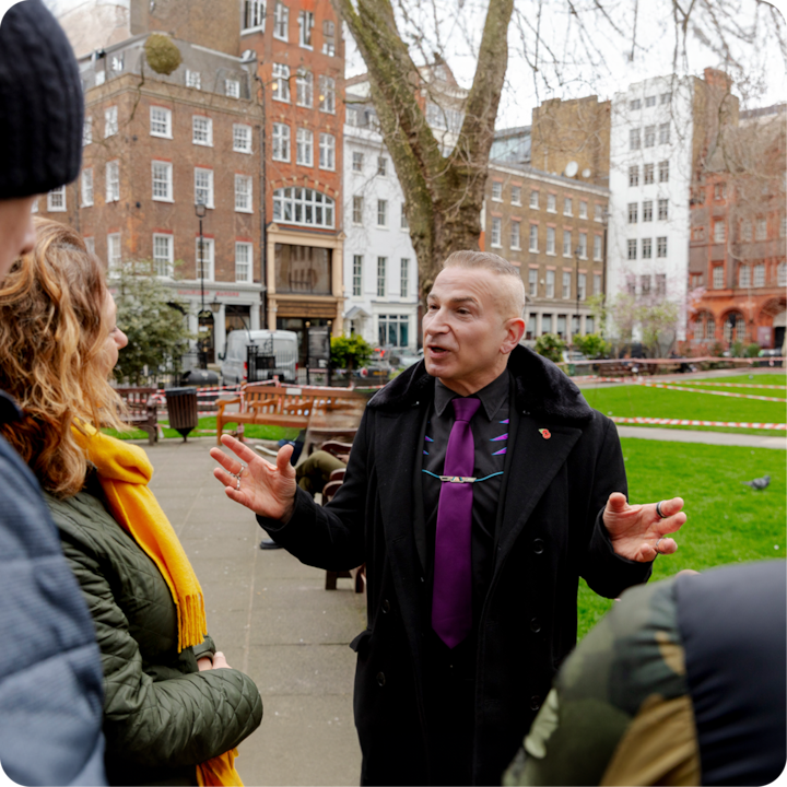 Un guide touristique vêtu d'un manteau noir et d'une cravate violette fait des gestes avec enthousiasme en parlant à un petit groupe sur une place historique avec des bancs et des arbres, entourée de bâtiments blancs et en briques.