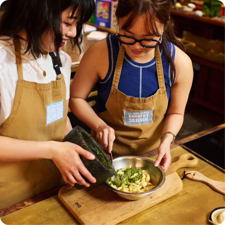 Deux participantes vêtues de tabliers bruns portant l'inscription « Cookery School » préparent des pâtes fraîches avec du pesto vert dans le cadre d'un atelier cuisine.