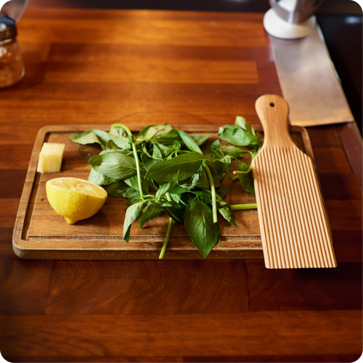 Une planche à découper en bois avec des feuilles de basilic, un demi-citron, un morceau de fromage et un ustensile en bois strié, posés sur une table en bois.