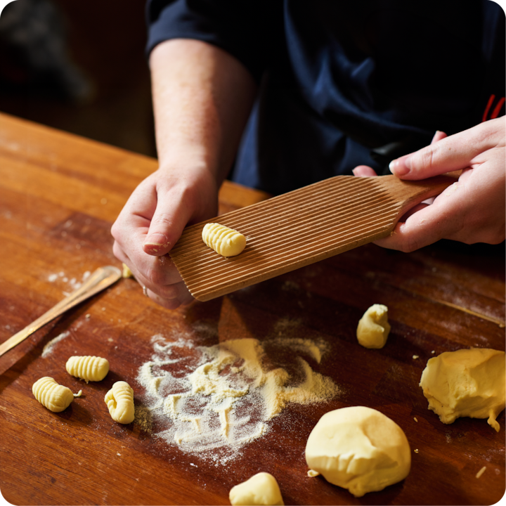 Des mains façonnent de la pâte à gnocchi fraîche sur une planche en bois striée. De la farine et des morceaux de pâte sont posés sur une table en bois.