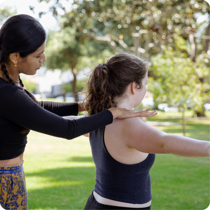 Une instructrice accompagne la position des bras d'un participant lors d'une séance de fitness en plein air dans un parc ensoleillé avec des arbres en arrière-plan.