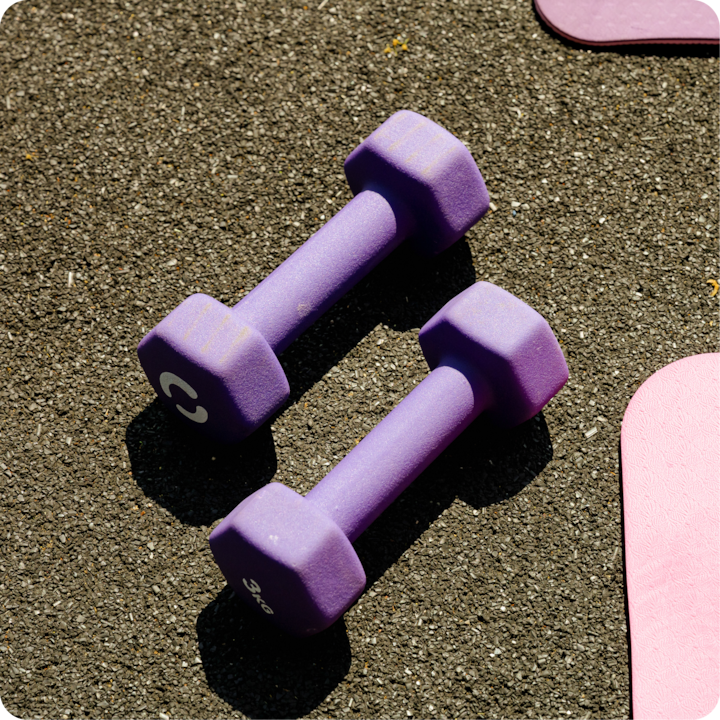Deux haltères violets sur de l'asphalte noir, avec le bord d'un tapis de yoga rose partiellement visible sur la droite.
