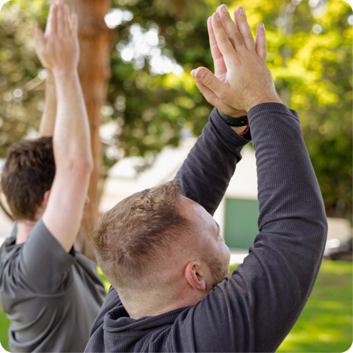 Dans un parc arboré, deux personnes réalisent une posture de yoga, les bras levés au-dessus de la tête.