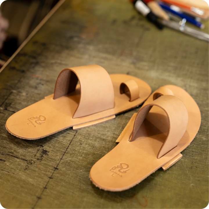 Plan rapproché de sandales en cuir fauve à brides incurvées, posées sur un établi et entourées d'outils d'artisan.