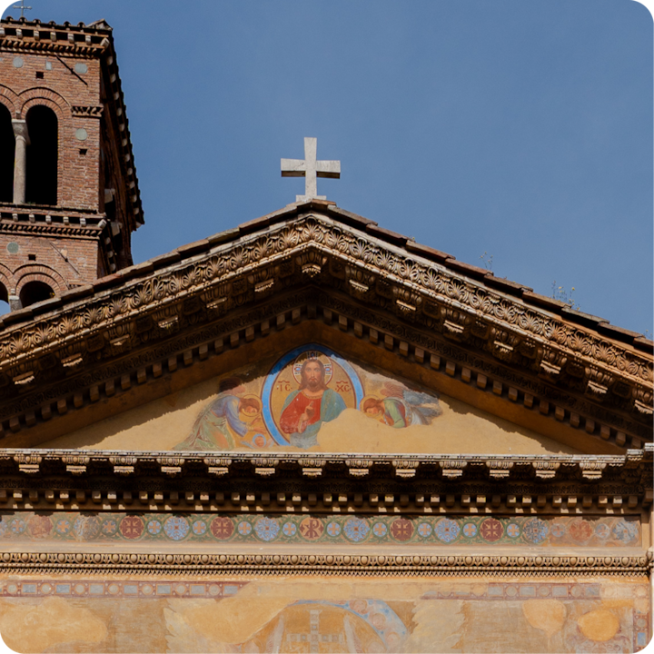 Façade d'église décorative avec un fronton triangulaire orné de fresques religieuses colorées, une croix en pierre au sommet et un clocher en briques sous un ciel bleu.