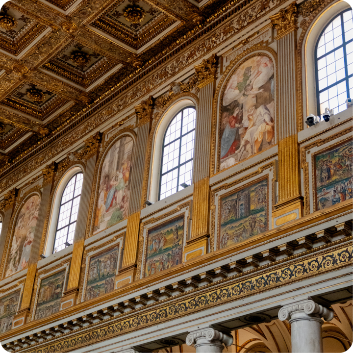 Intérieur décoratif d'un bâtiment historique avec de grandes fenêtres cintrées, des touches dorées et des fresques détaillées représentant des scènes historiques et religieuses.