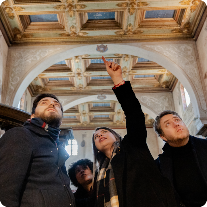 Un groupe de personnes se tient à l'intérieur d'un bâtiment historique avec des arches décorées et un plafond peint, tandis qu'une personne fait un geste vers le haut, guidant une visite.