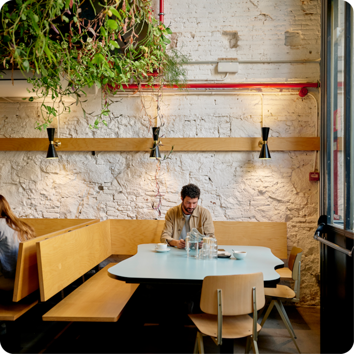 Un café confortable avec des meubles modernes en bois, des plantes suspendues et un éclairage de style industriel, montrant un coin salon lumineux avec une table dressée pour une personne.