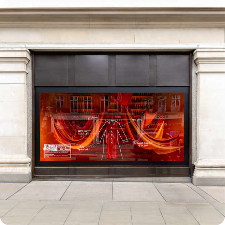 Une vitrine de magasin avec un design rouge et orange vif, des rideaux en tissu, le croquis d'une tenue rouge et des détails étiquetés, avec des reflets de la rue dans la vitre.