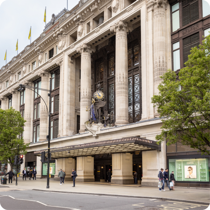 Un bâtiment historique avec de grandes colonnes et une sculpture en forme de globe au-dessus de l'entrée, situé dans une rue animée bordée d'arbres où circulent des piétons.