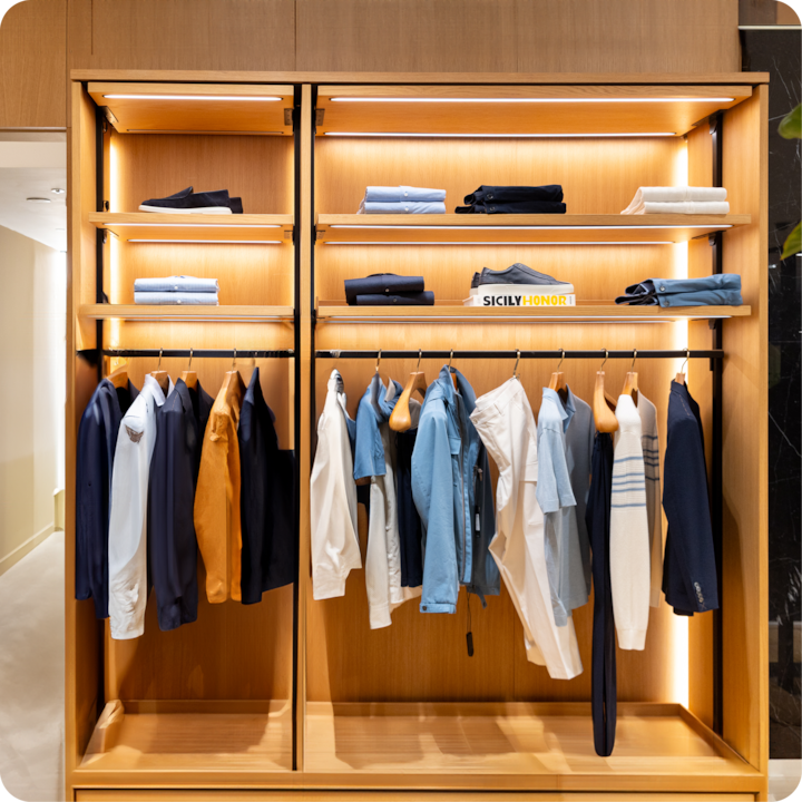 Une garde-robe en bois baignée d'une lumière douce contenant des vêtements pliés soigneusement disposés, des chemises suspendues dans des tons de bleu, de blanc et de beige, ainsi qu'une paire de chaussures et un livre intitulé « Sicily Honor » sur les étagères.