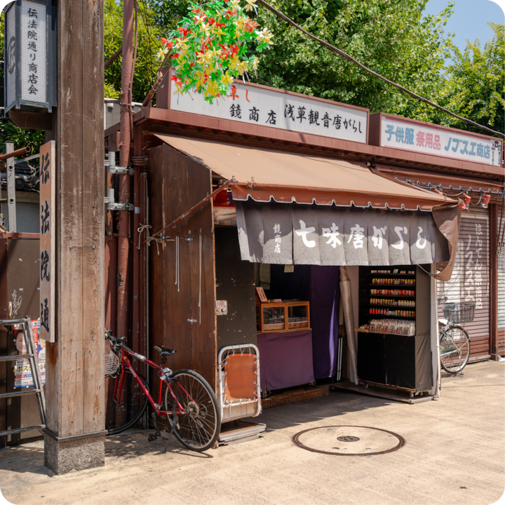 Un étal de marché en plein air au design traditionnel japonais, présentant des produits colorés et un vélo rouge appuyé contre un poteau en bois, sous un ciel bleu.