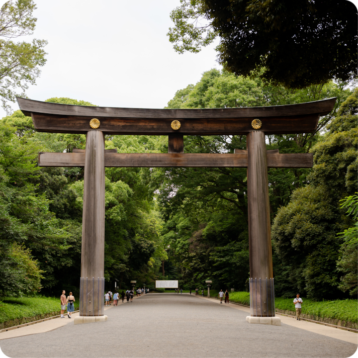 Un portail torii japonais traditionnel aux accents dorés se dresse dans un paysage de forêt serein, avec des visiteurs qui se promènent sur le sentier en contrebas et une végétation dense en arrière-plan.
