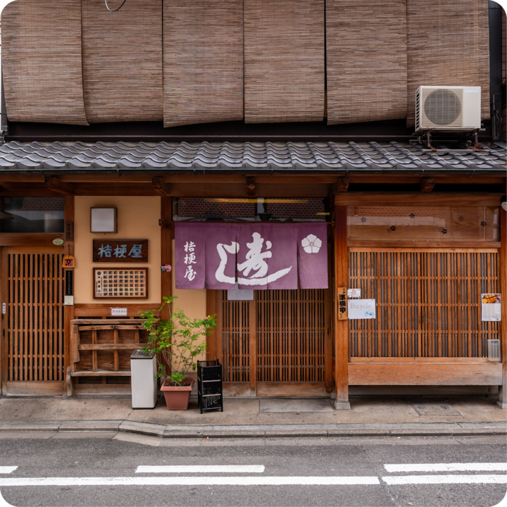 L'extérieur d'un bâtiment japonais traditionnel avec des détails en bois, un noren violet orné de kanjis et de petites plantes en pot près de l'entrée.