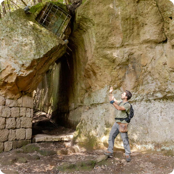 Un cadre extérieur rural avec d'anciennes structures en pierre, un passage rocheux et une lumière naturelle filtrant à travers les arbres, où une personne explore le site.