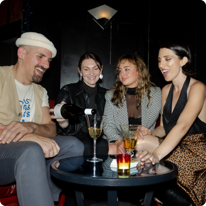 Plan rapproché de personnes réunies autour d'une table dans un salon chic et sombre, éclairées par un flash. Une bouteille de vin est versée dans un verre entouré d'autres boissons et d'une petite bougie.