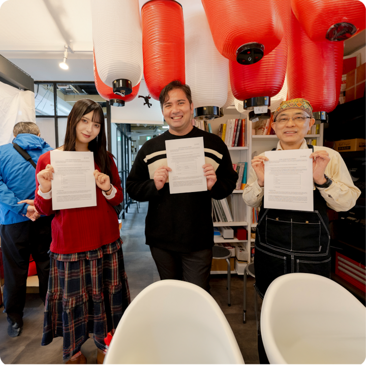 Trois personnes se tiennent en intérieur, tenant des formulaires de décharge signés. Des lanternes rouges et blanches sont suspendues et des étagères remplies de livres et de fournitures se trouvent en arrière-plan.