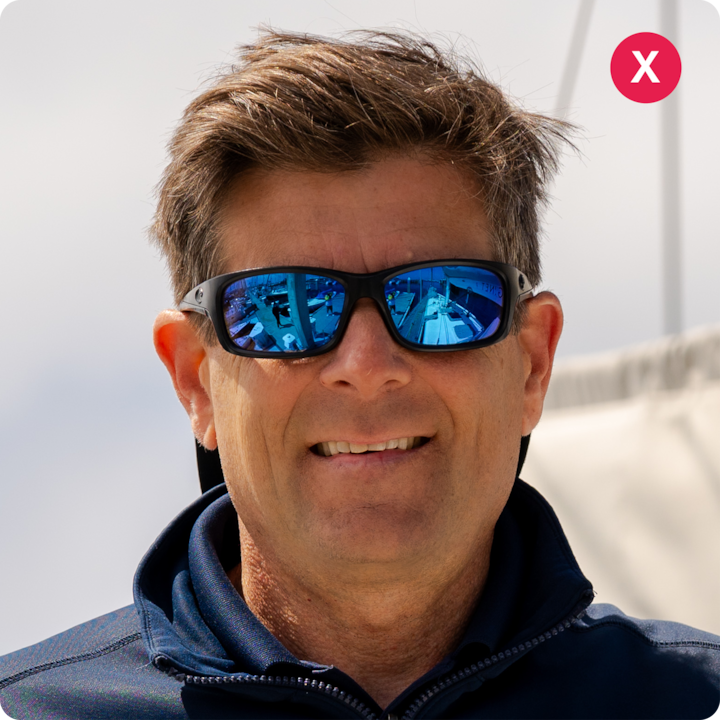 Plan rapproché d'une personne portant des lunettes de soleil et une veste bleu marine, debout à l'extérieur, avec une partie d'un voilier et un ciel nuageux en arrière-plan.