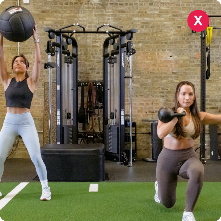 Deux femmes s'entraînent dans une salle de sport. L'une lance un wall ball au-dessus d'elle et l'autre fait une fente avec un kettlebell sur un sol effet gazon. Des équipements de sport et un mur de briques sont visibles en arrière-plan.
