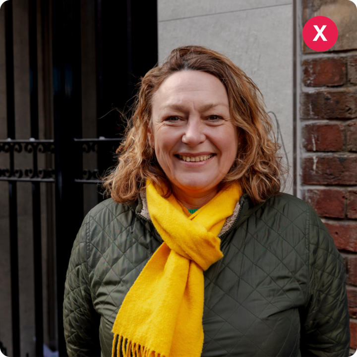 Une voyageuse portant une veste matelassée verte et une écharpe jaune vif se tient devant un mur de briques et une porte en métal noir.