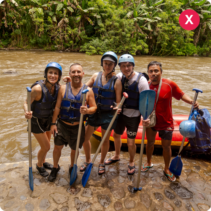 Un groupe de cinq personnes portant des gilets de sauvetage et des casques, tenant des pagaies, se tient sur un chemin de pierre à côté d'un canoë gonflable jaune et rouge au bord d'une rivière. Une jungle verdoyante les entoure.