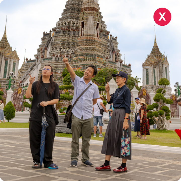 Trois personnes se tiennent debout devant le temple Wat Arun à Bangkok, en Thaïlande, pointant du doigt vers le haut. De la verdure bien entretenue et d'autres visiteurs se trouvent en arrière-plan.