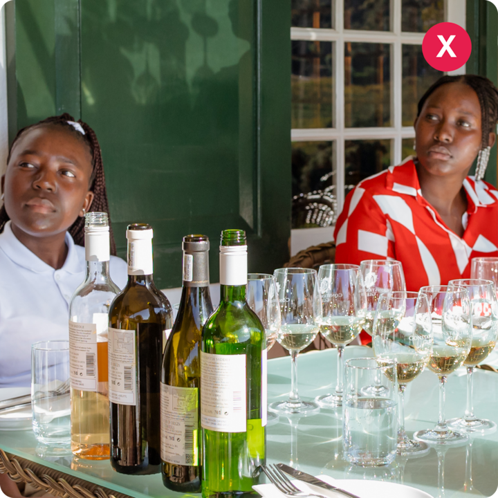 Une table dressée avec plusieurs bouteilles de vin, des verres à vin vides et des verres à eau dans un cadre extérieur, avec des murs peints en vert et une fenêtre reflétant de la verdure en arrière-plan.