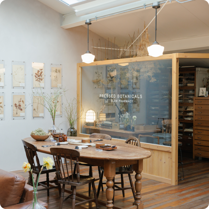 Une pièce accueillante avec une table en bois entourée de chaises, préparée pour une activité de création d'herbiers. Derrière la table, un mur de verre porte l'inscription « Pressed Botanicals - Slow Pharmacy », avec un décor naturel et des plantes séchées qui mettent l'atmosphère en valeur.