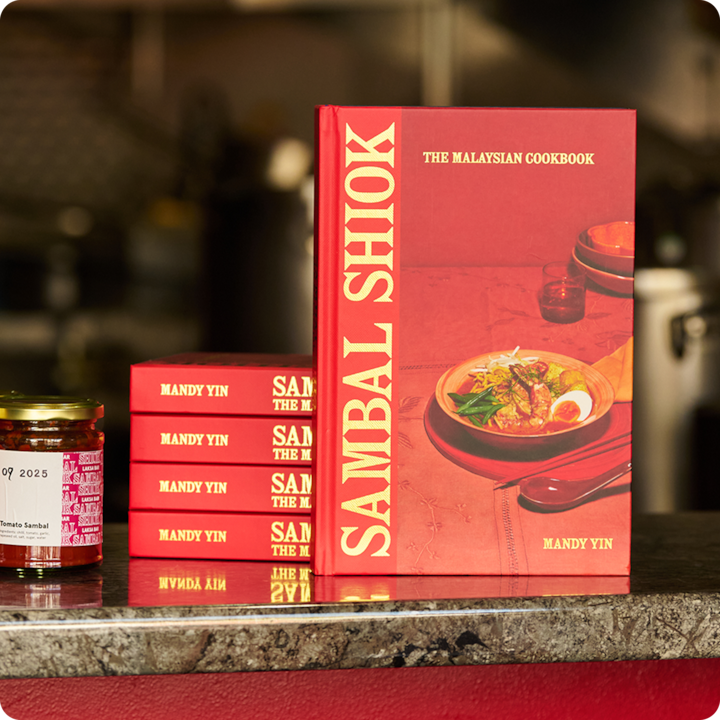 Une pile de livres de cuisine rouges sur un plan de travail, à côté d'un bocal. Un bol de nourriture malaisienne est représenté sur la couverture du livre.