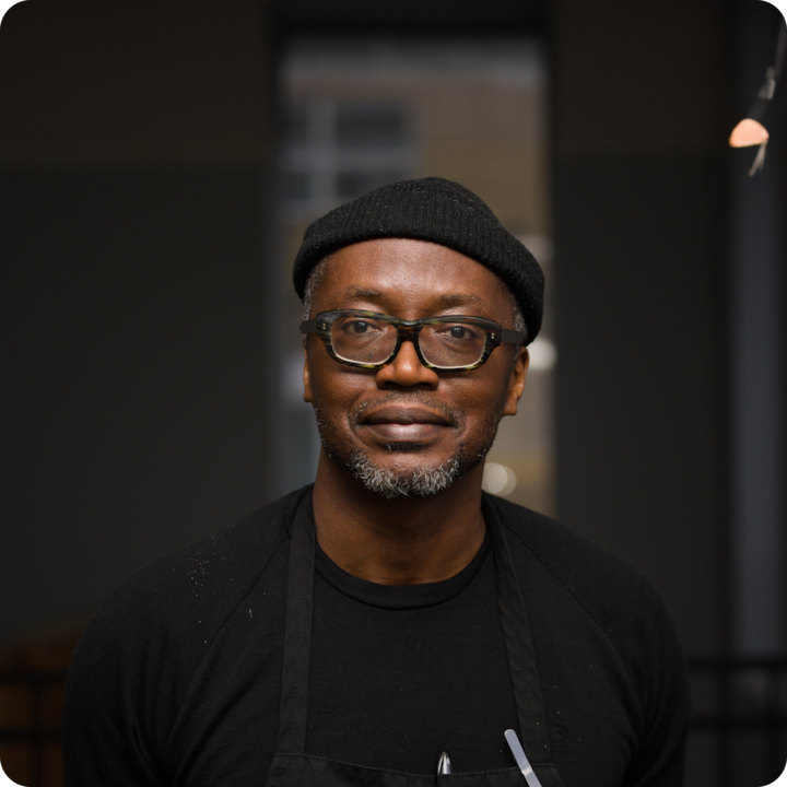 Sous un éclairage chaleureux, une personne portant un bonnet noir, un tablier noir et un t-shirt noir se tient debout à l'intérieur sur fond de fenêtres flou.