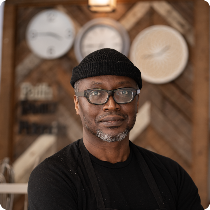 Une personne portant un tablier noir et un t-shirt noir se tient dans un cadre rustique et chaleureux avec un mur en bois orné d'horloges et de panneaux décoratifs en arrière-plan.