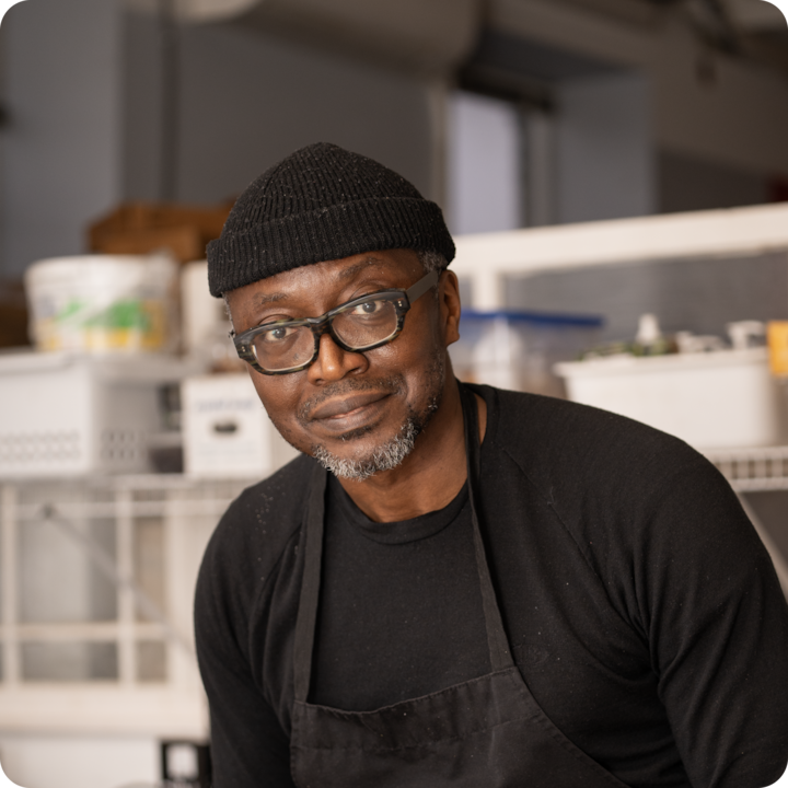 Une personne portant un bonnet noir et un tablier se tient dans une cuisine accueillante avec des étagères contenant des récipients et des articles de cuisine en arrière-plan.