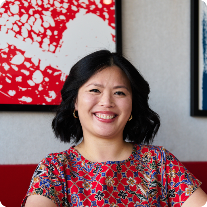 Une personne vêtue d'un chemisier rouge à motifs floraux colorés est assise à l'intérieur. Des œuvres d'art abstraites rouge et blanc encadrées se trouvent sur le mur derrière elle.