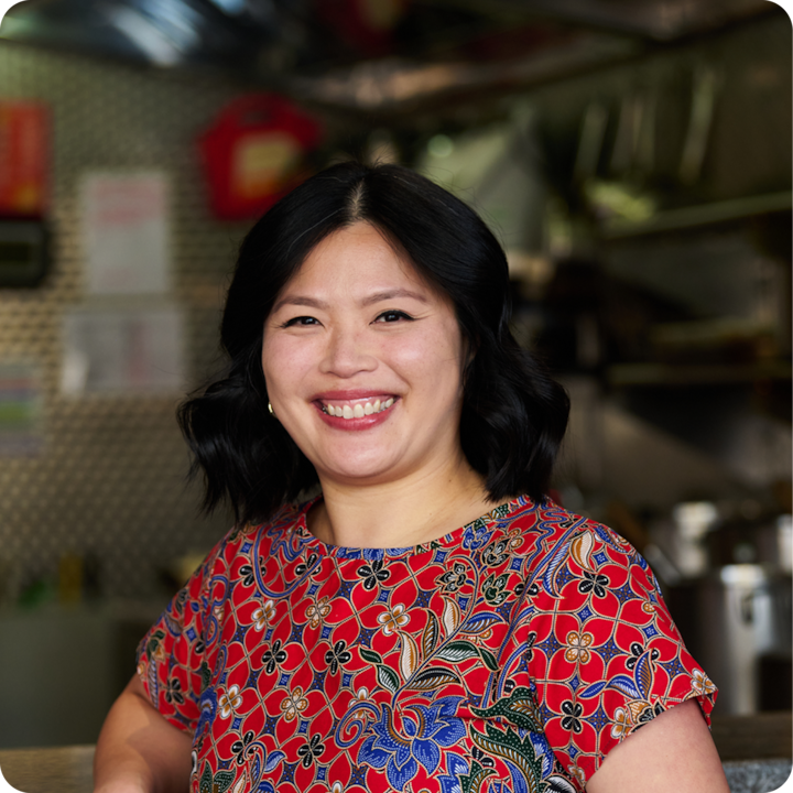 Une personne portant un chemisier rouge vif à motifs floraux se tient dans une cuisine chaleureuse avec des murs métalliques et des ustensiles de cuisine en arrière-plan.