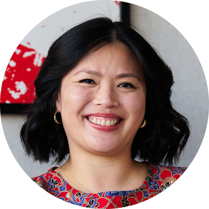 Une personne aux cheveux noirs mi-longs portant un haut rouge à motifs, le visage flou, se tient debout en intérieur contre un mur de couleur claire. Une œuvre d'art abstraite rouge est visible en arrière-plan.