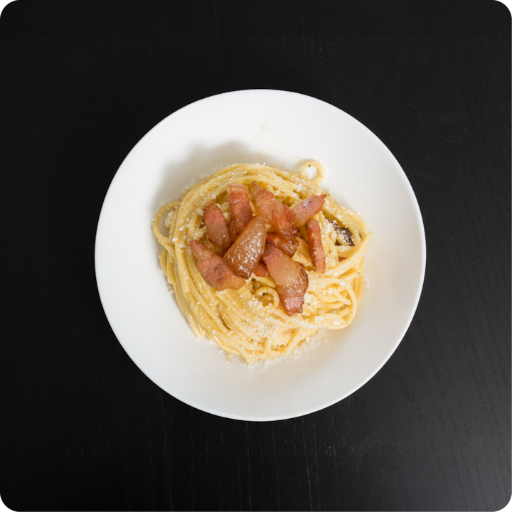 Une assiette de spaghettis carbonara garnie de lardons croustillants et d'une pincée de fromage râpé, posée sur une table noire.