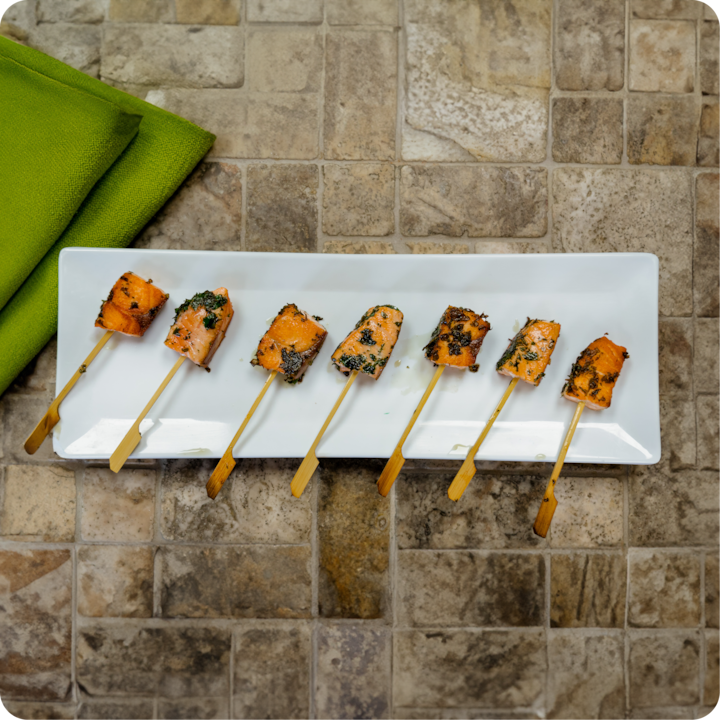 Une assiette blanche rectangulaire contient sept morceaux de saumon grillé aux herbes sur des brochettes de bambou. Elle est posée sur une surface carrelée texturée avec des serviettes vertes pliées à proximité.