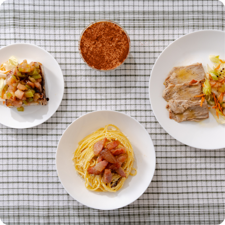 Une table avec trois assiettes blanches présentant des plats : des spaghettis au bacon, une pâte garnie de légumes et de la viande en tranches avec une salade fraîche, autour d'un petit bol de tiramisu sur une nappe à carreaux.