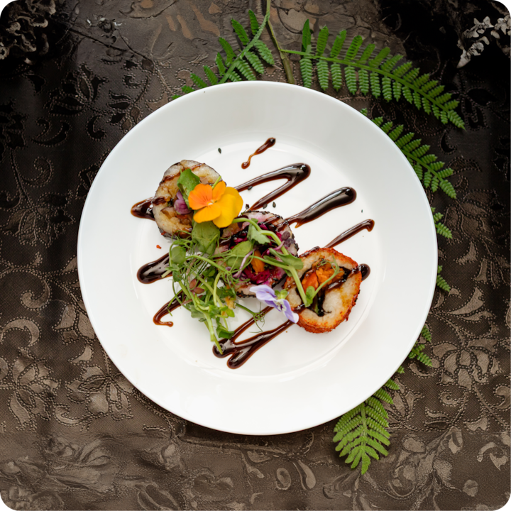 Trois rouleaux de sushi garnis de fleurs et d'herbes comestibles colorés, élégamment disposés sur une assiette blanche avec un filet de vinaigre balsamique, placés sur une nappe sombre texturée avec des accents de fougères.