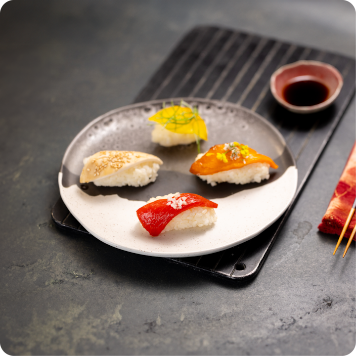 Assiette de quatre sushis colorés, dont un au poivron rouge, un à la mangue jaune, un au saumon orange et un blanc garni de sésame, servis avec de la sauce soja.